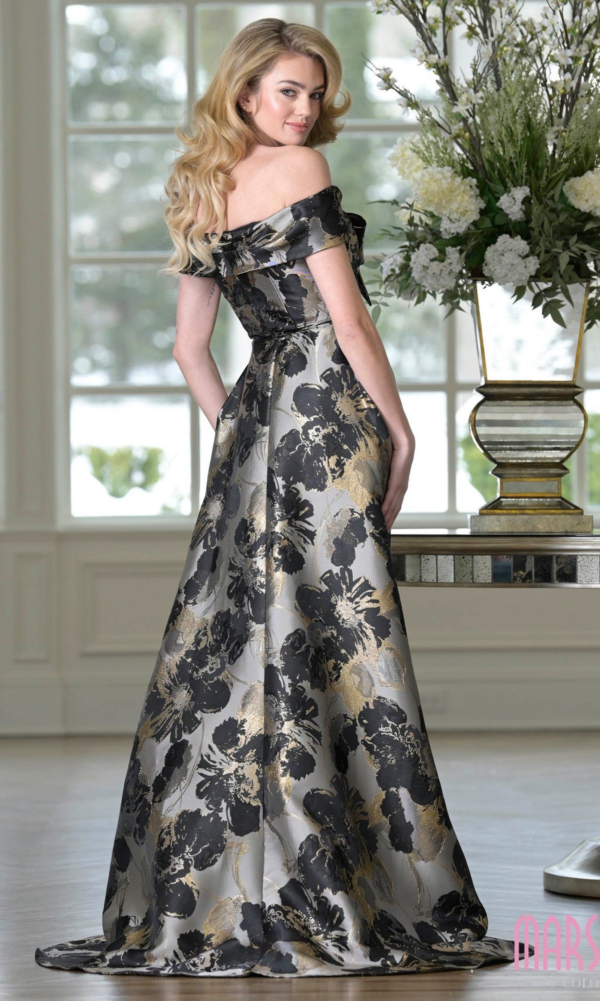 A woman stands indoors by a table with a large floral arrangement, wearing the Marsoni MV1396—a long formal off-the-shoulder gown in black and gold jacquard, perfect for a mother-of-the-bride look.